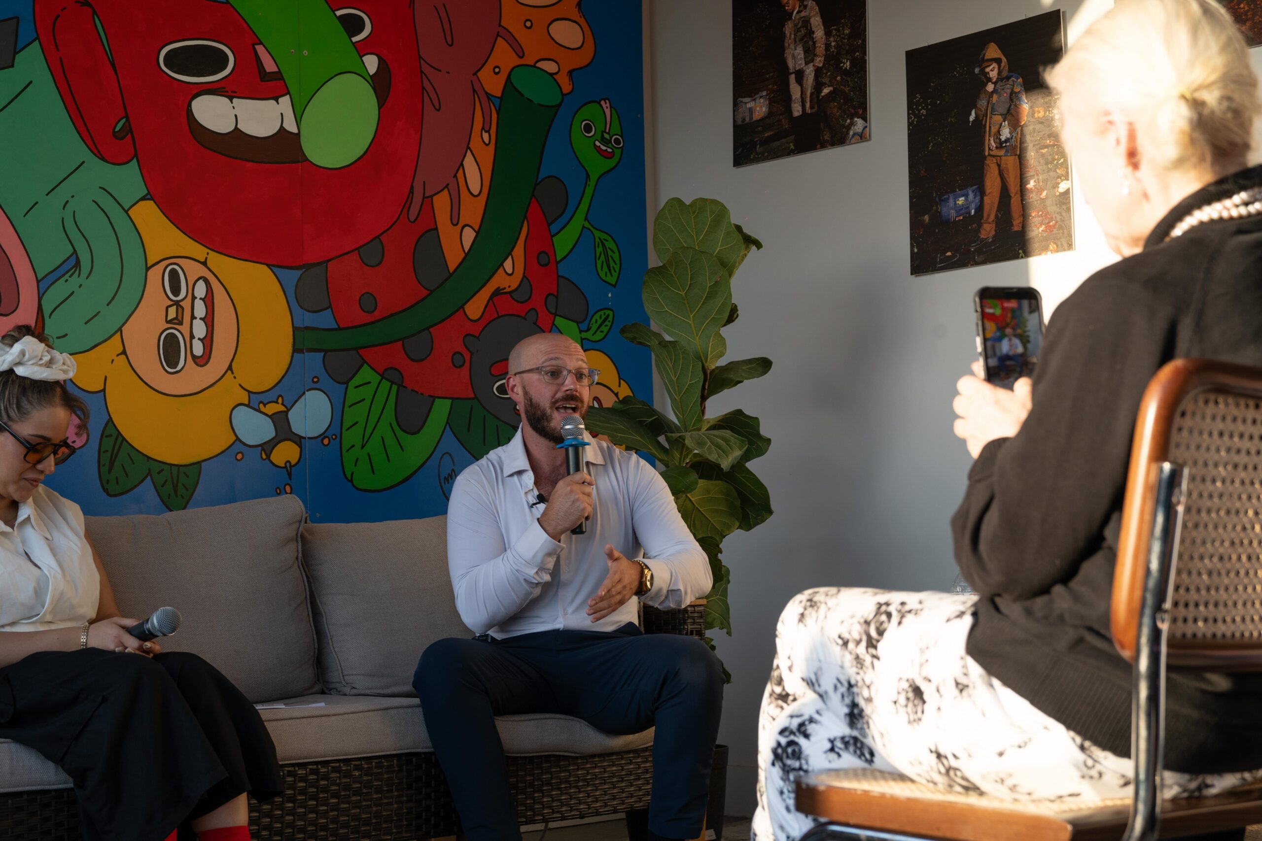 A person speaks into a microphone while seated on a couch, with another person beside them and someone in the foreground recording with a phone. Colorful mural art is on the wall behind.