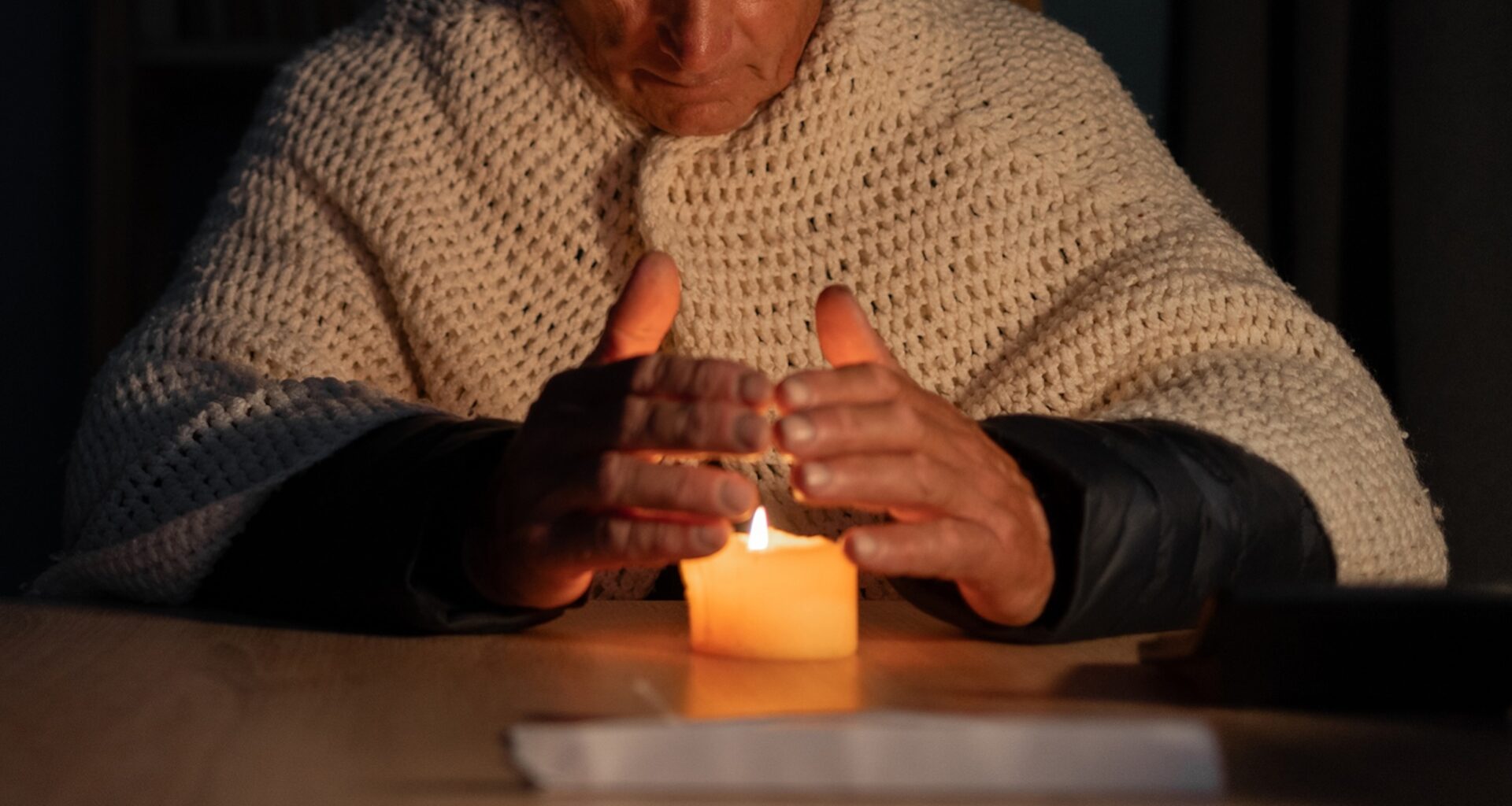 A senior citizen sitting at a table with a lit candle, hands placed above it, wearing a knitted blanket.