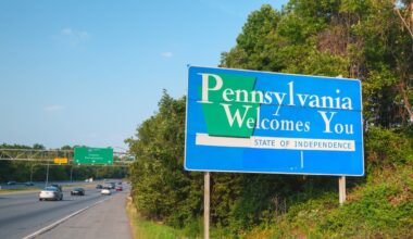 A blue 'Pennsylvania Welcomes You' sign with green accents stands on the right side of a multi-lane highway. In the background, an overhead green sign for I-95 North points to Chester and Philadelphia. Several cars are driving on the highway, and green trees line both sides of the road under a clear blue sky.