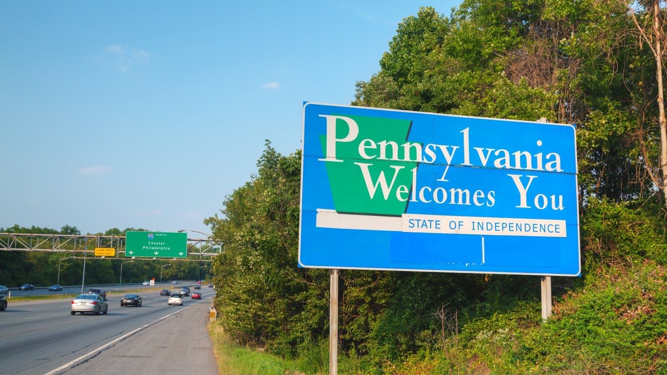 A blue 'Pennsylvania Welcomes You' sign with green accents stands on the right side of a multi-lane highway. In the background, an overhead green sign for I-95 North points to Chester and Philadelphia. Several cars are driving on the highway, and green trees line both sides of the road under a clear blue sky.