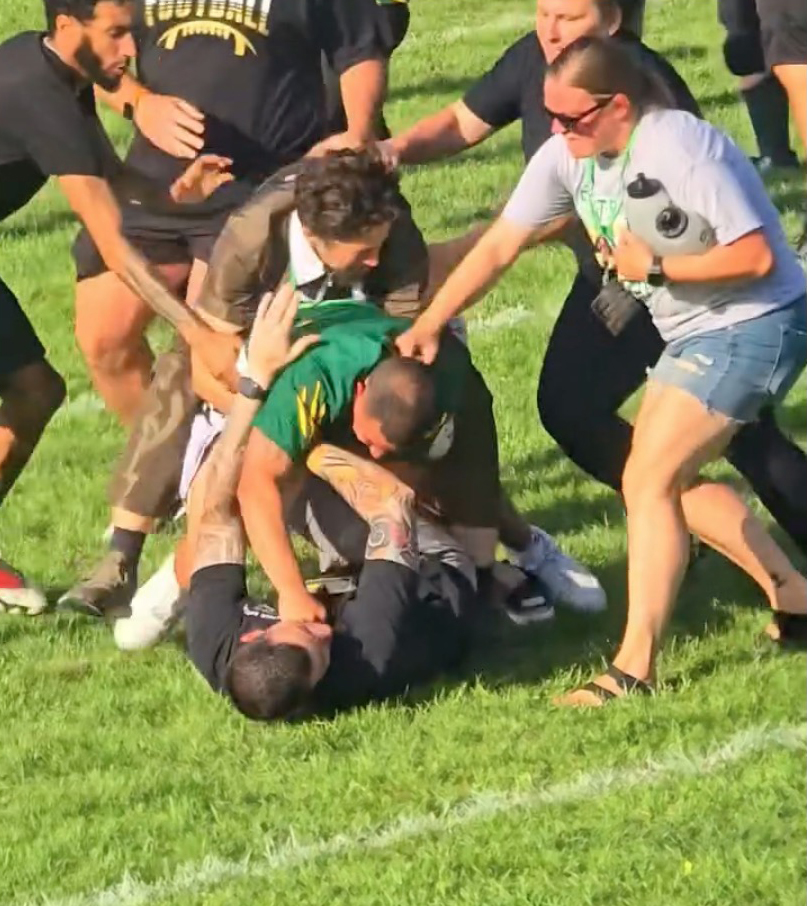 A youth football coach in a black shirt being tackled to the ground by another man, while several adults attempt to break up the fight on a grassy field.