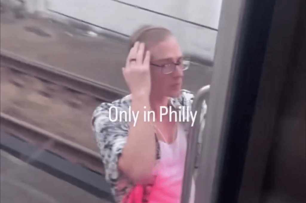 A man in glasses, a patterned shirt, and a pink tank top hangs from the side of a moving SEPTA train, with "Only in Philly" superimposed on the image.