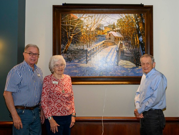 Kirkland Village Art Commission members Margie Mosher, Ned Bowne, and Charles Lyman stand near a painting of Bethlehem's 1754 Waterworks by Sergei Yaralov, one of four recently installed in the main dining room of Kirkland Village, on Friday, Oct. 2, 2026, in Bethlehem. Residents donated money to purchase the art, part of a collection of over 800 pieces. The 1754 Waterworks pumped spring water up to a water tower that stood where Central Church stands today, from which the water flowed by gravity to town buildings. The restored building, containing a waterwheel and a three-piston pump, is a National Historic Landmark and is known as the first municipal pumped water system in the United States. (April Gamiz/The Morning Call)