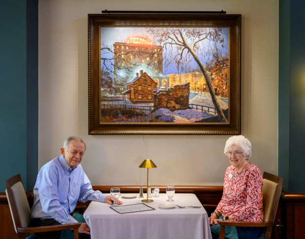 Kirkland Village Art Commission members Charles Lyman and Margie Mosher sit under a painting of The 1750 Smithy by Sergei Yaralov, one of four recently installed in the main dining room of Kirkland Village, on Friday, Oct. 2, 2026, in Bethlehem.  Residents donated money to purchase the art, part of a collection of over 800 pieces. The 1750 Smithy was reconstructed in 2004 from archeological evidence and an original drawing that has been preserved in the Moravian Archives. Trades housed in this building were blacksmithing, locksmithing, nailsmithing, and tinsmithing. Today, blacksmiths demonstrate the production of iron products. The free-standing wall in the foreground is all that remains of the pottery. (April Gamiz/The Morning Call)