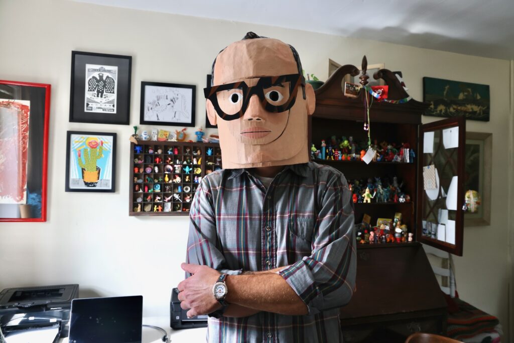 James Heimer is in his home, wearing an oversized cartoonish mask that he madeof his own face.