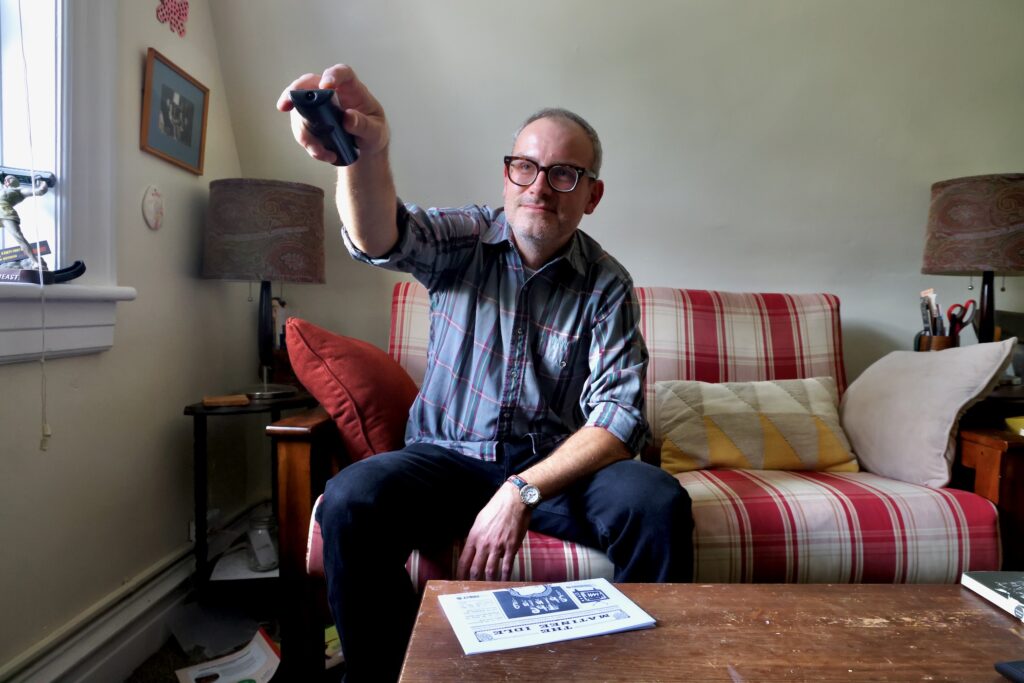 James Heimer is sitting on the couch in his home pointing the remote control at the television.