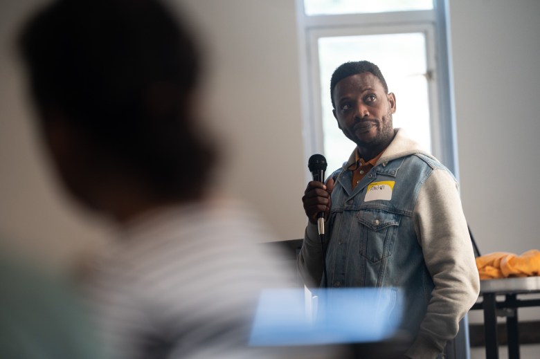 A man with a microphone and a name tag stands indoors, looking to the side, with another person blurred in the foreground.