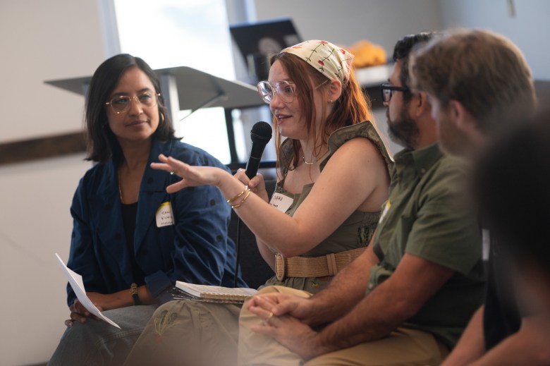 Four people sit in a row at a panel discussion; one woman with a headscarf speaks into a microphone while others listen.