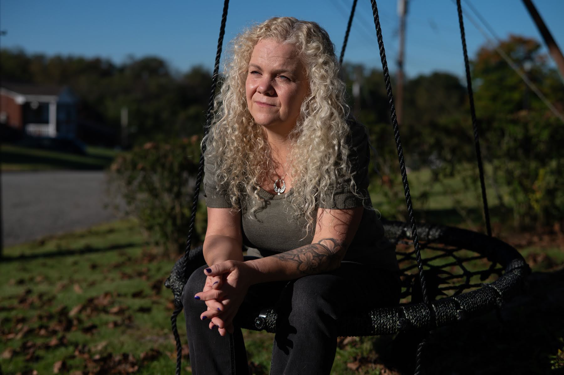 A woman with long curly blonde hair sits on a black saucer swing in a yard.