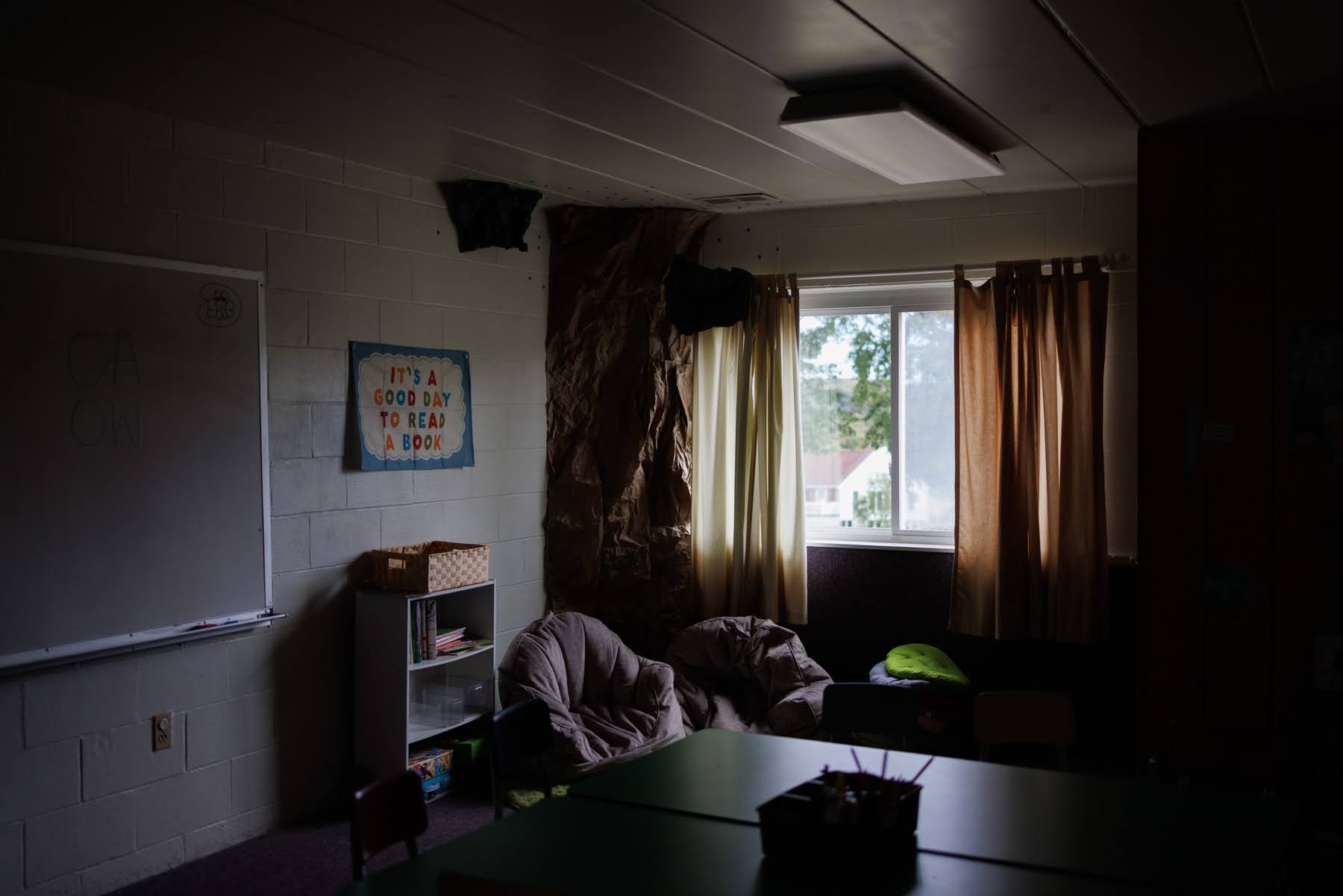 A dimly lit classroom corner with a whiteboard, a poster that says “It’s a good day to read a book,” a small bookshelf, and two beanbag chairs near a window.
