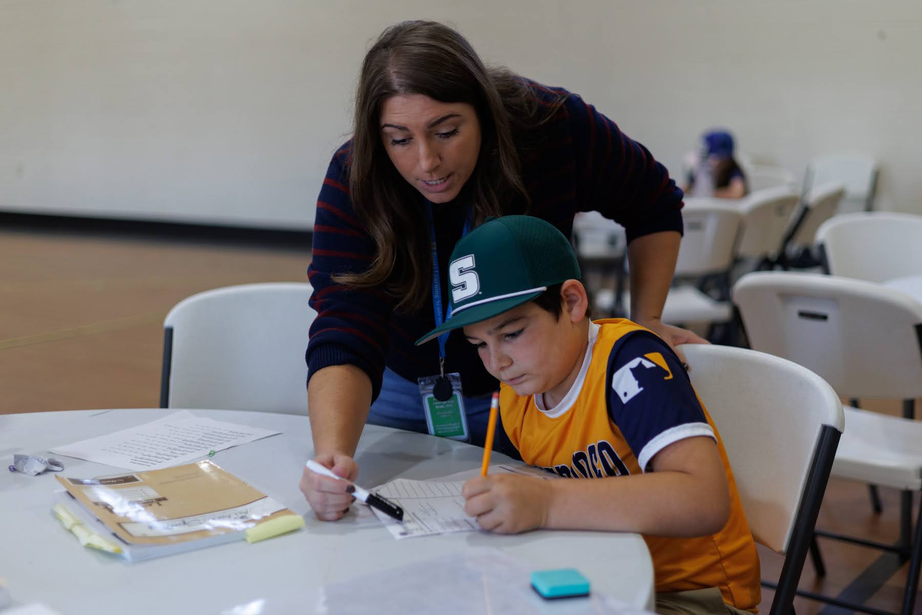 A teacher leans over a table to help a young boy in a baseball cap write on a paper.