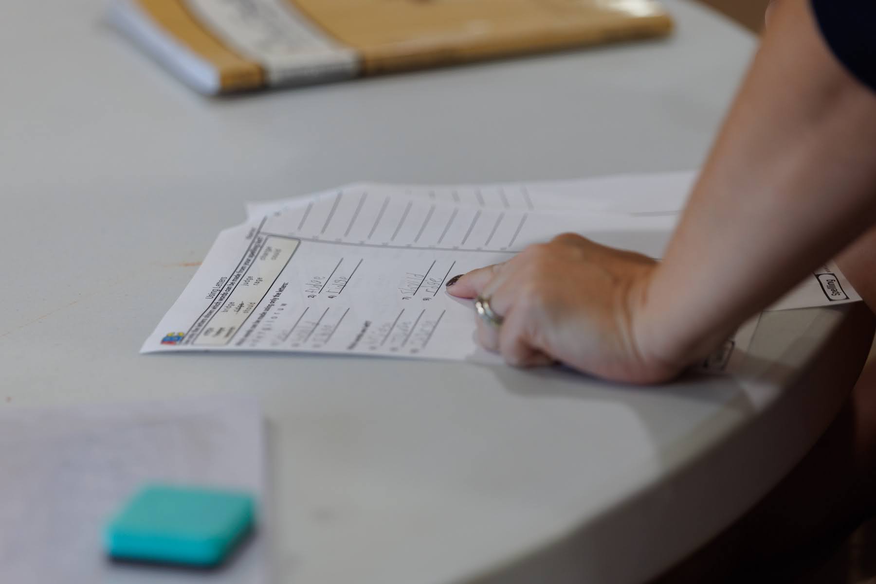 A hand points to a handwritten spelling worksheet on a table.