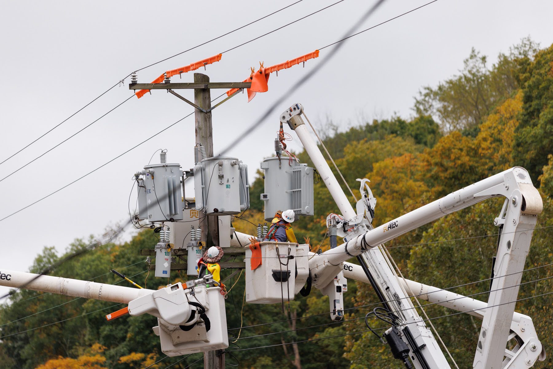 Two utility workers in bucket trucks perform maintenance on electrical transformers attached to a wooden utility pole.