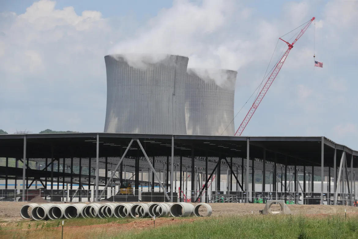 A metal building is being constructed, with water cooling towers loom in the background.