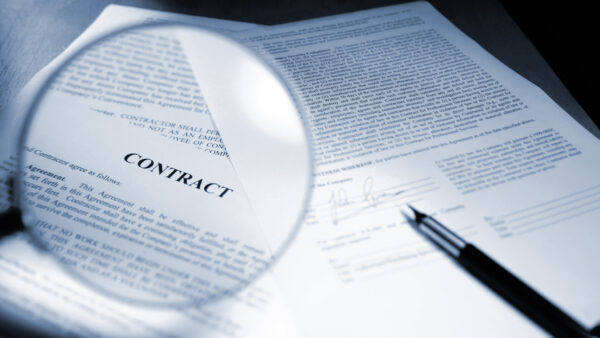 A close up blue toned image of a magnifying glass held out in front of page in a legal contract document. Sheets of paperwork are on a wooden desk with a ink pen close to a signed page. Contract text and signature are fake. Shot with shallow depth of field with focus on the word ‘Contract’.
