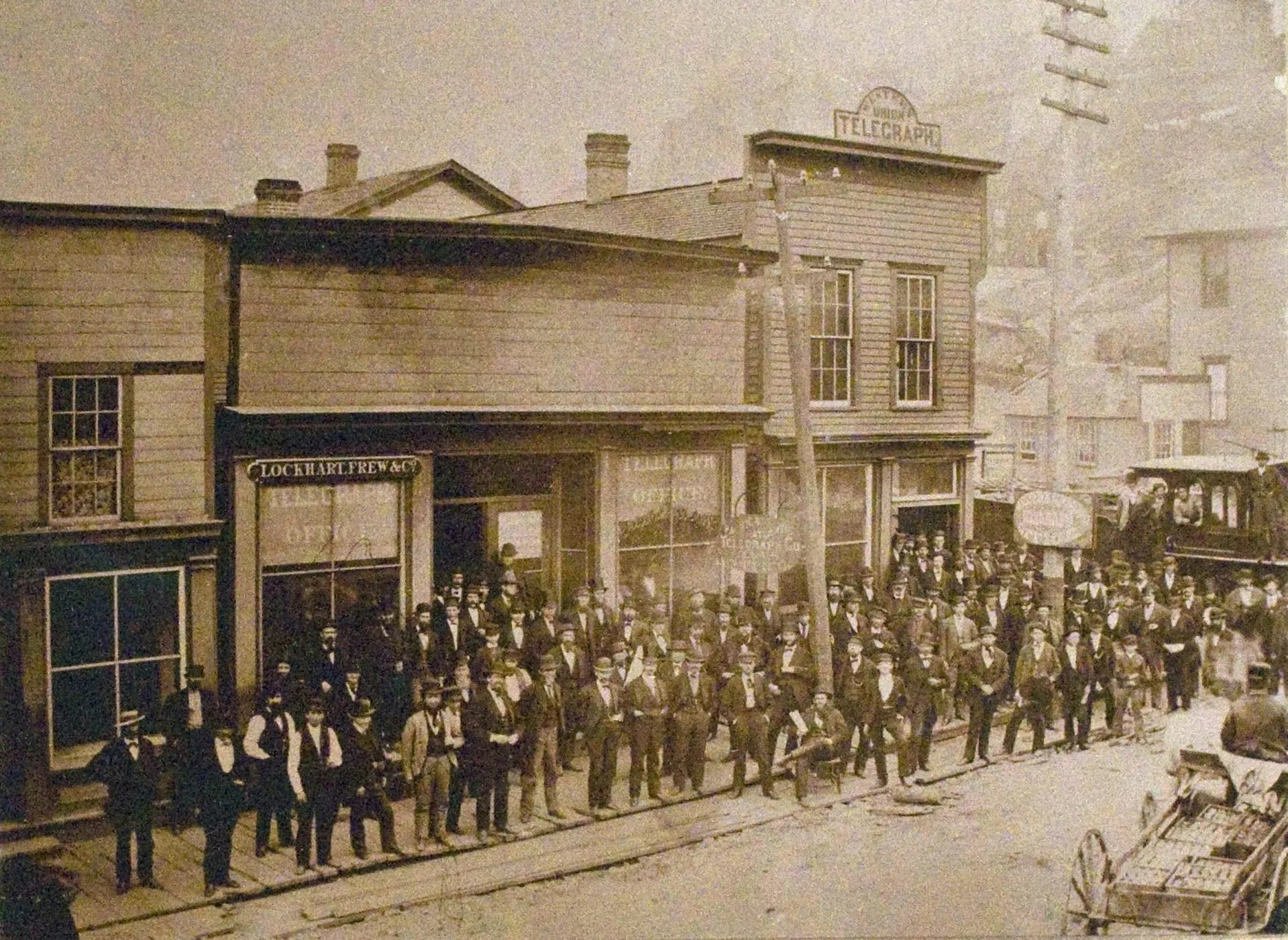 Dozens of men in suits stand along a curb in a sepia-toned photo.
