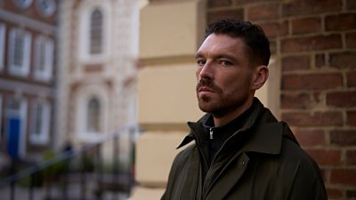 A man in green jacket and stood against a brick wall