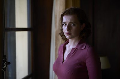 A woman in a red jumper staring intently into the camera next to a window of a grand room