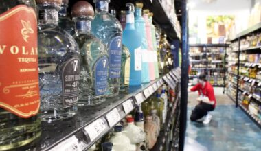 Bottles of tequila on a shelf in a store with a worker wearing a red top and black pants stocking inventory in the background.
