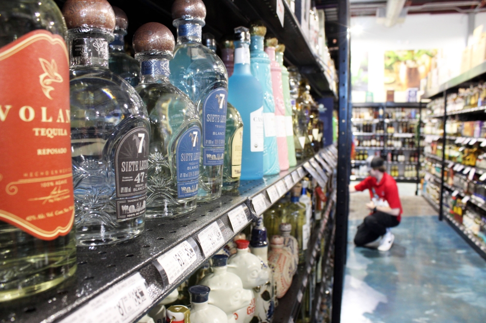 Bottles of tequila on a shelf in a store with a worker wearing a red top and black pants stocking inventory in the background.