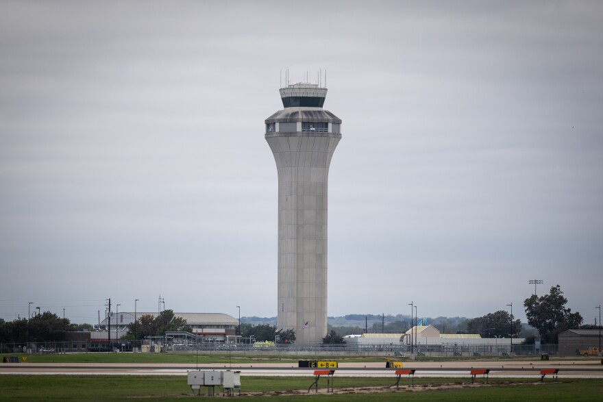 The air traffic control tower of Austin-Bergstrom International Airport, a large grey concrete tower surrounded by a security fence against the backdrop of an overcast sky. On the ground, various airport infrastructure is visible including a two story building.  