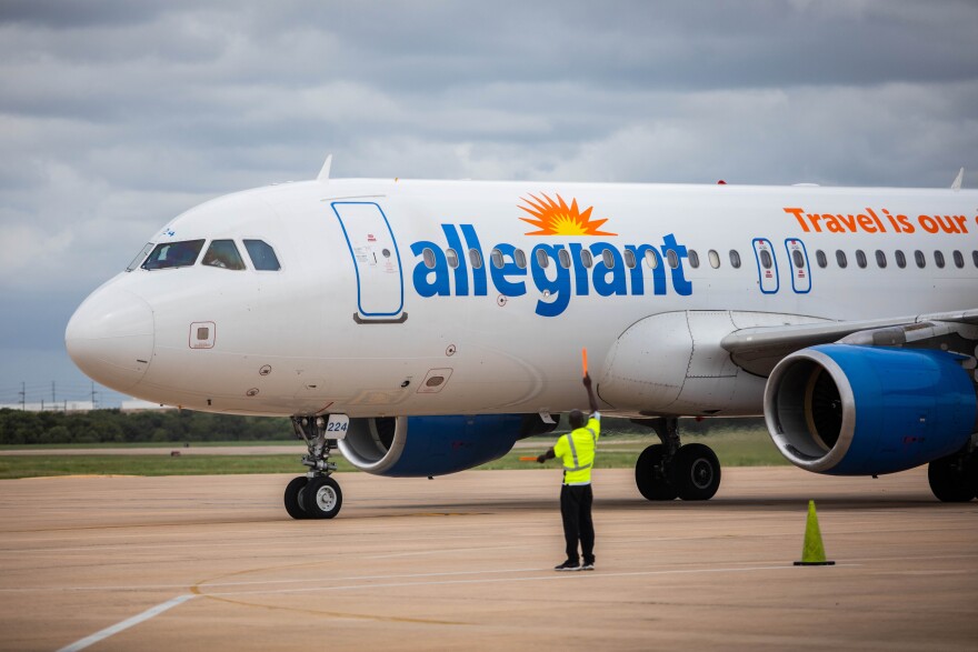 A ground crew member directs an Allegiant Air airplane on the tarmac at an airport. The plane features the Allegiant logo with a sunburst on its fuselage. The crew member is wearing a bright yellow safety vest and using orange wands to guide the aircraft. The sky is overcast, and there are industrial buildings visible in the background.