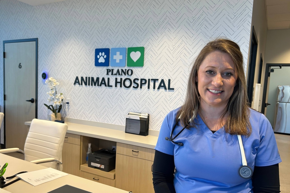 Female wearing medical scrubs and a stethoscope around her neck stands in front of a wall featuring a Plano Animal Hospital sign.