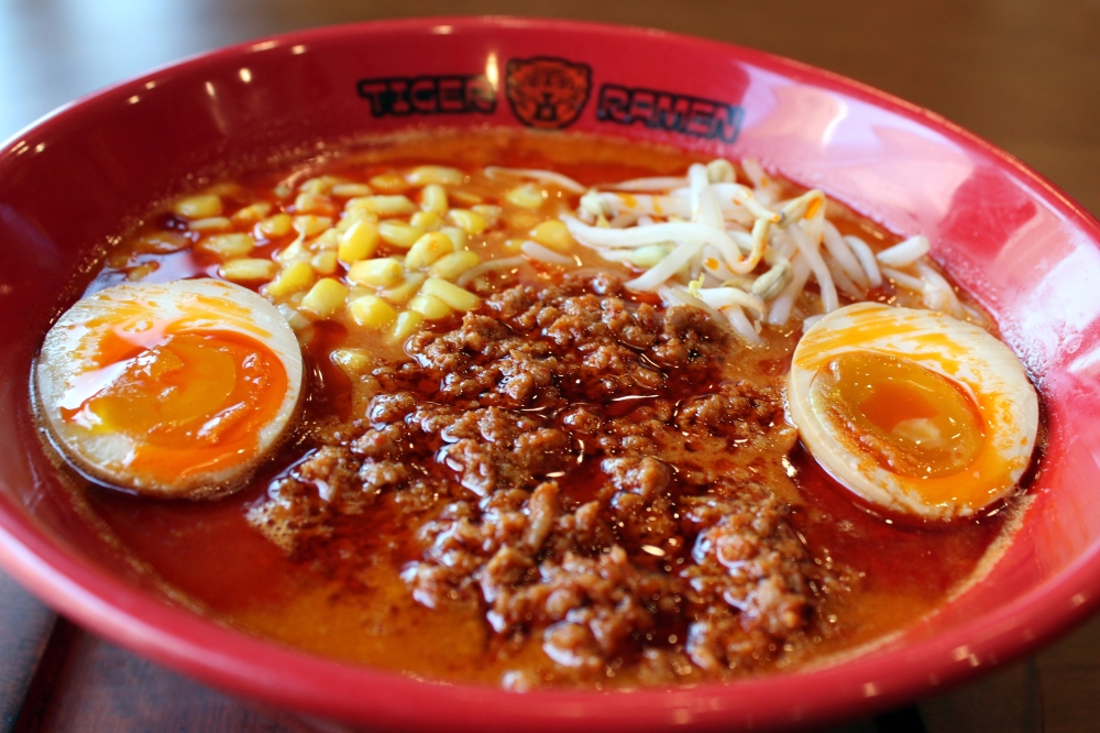 Red bowl featuring Tiger Ramen branding is filled with ramen, meat, eggs, corn and broth.