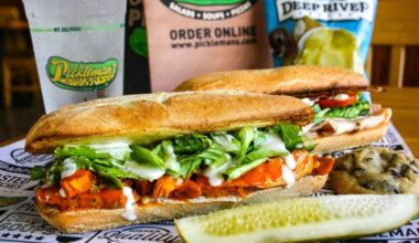 Sub sandwich with a side pickle in front of drinks featuring Pickleman's branded cups.