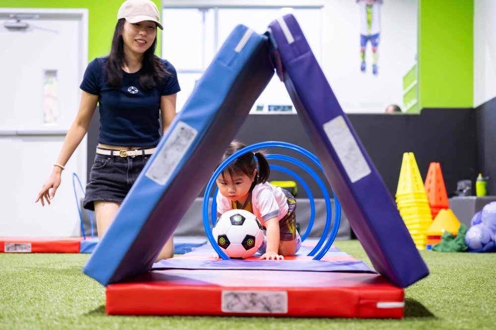 A toddler pushes a soccer ball through an obstacle course while a woman observes