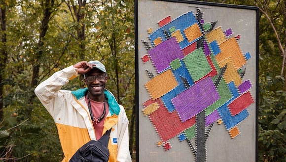 A person in outdoor clothing stands in a wooded setting with a standing public sculpture resembling a quilted tree in block colors of purple, red, green, yellow, and blue.