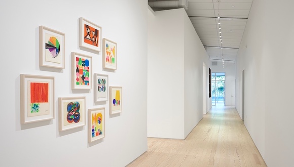 A view down the long hallway of an architecturally renovated gallery space, with framed artworks in the foreground and a window outside in the background.
