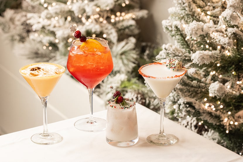 Four festive cocktails and mocktails are set on a table with a snow-flocked Christmas tree...