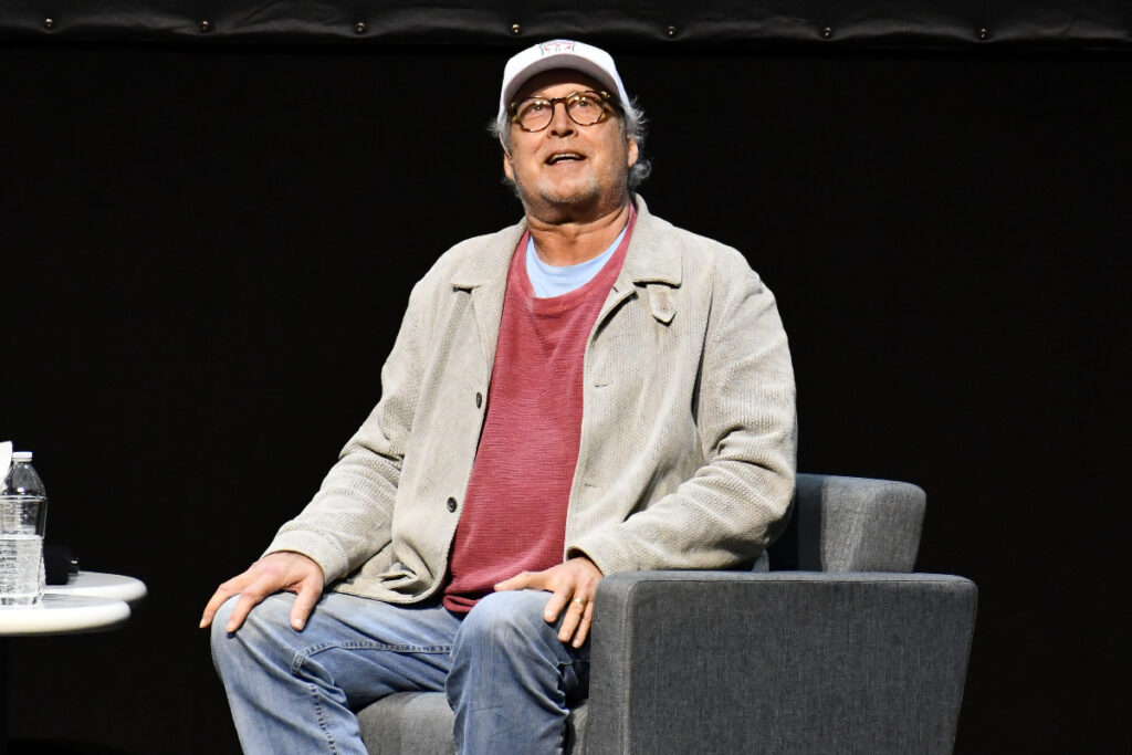 Image shows Chevy Chase sitting in a chair onstage for 