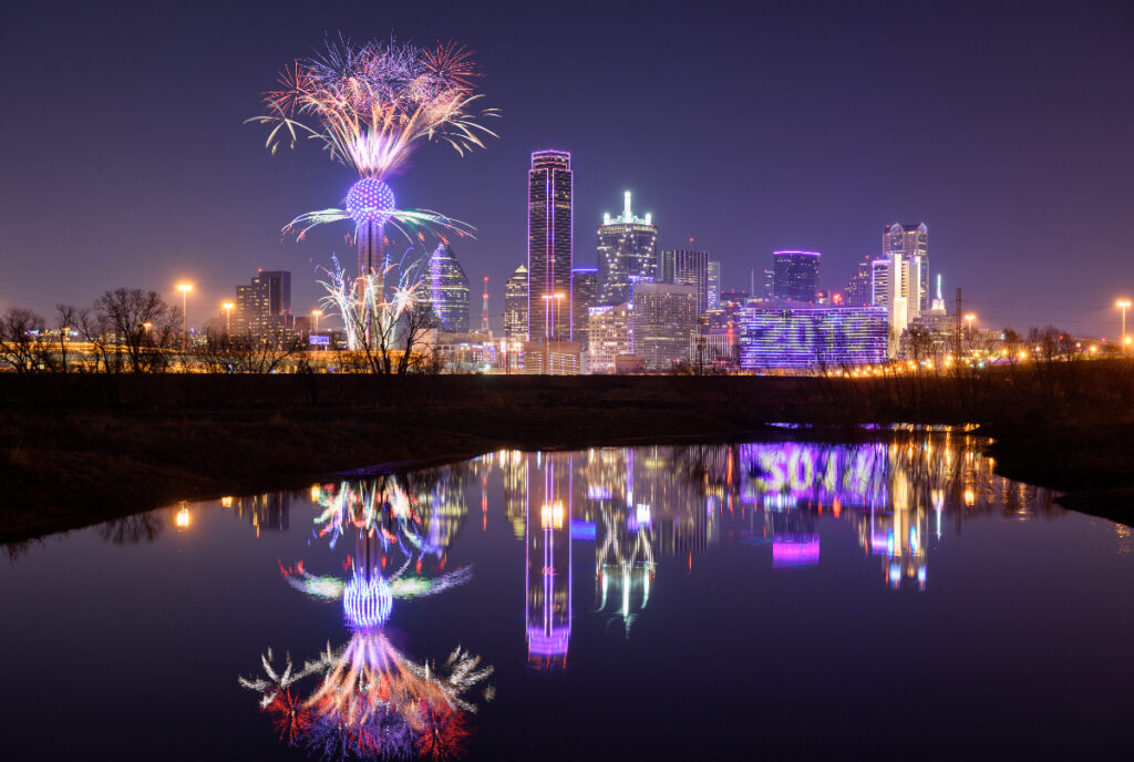 Image shows the fireworks at Reunion Tower in the Dallas skyline across a body of water.