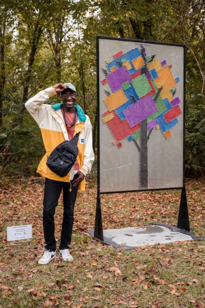 A person in outdoor clothing stands in a wooded setting with a standing public sculpture resembling a quilted tree in block colors of purple, red, green, yellow, and blue.
