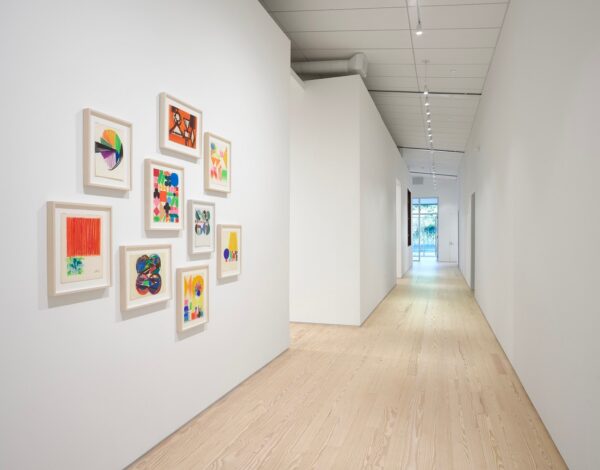 A view down the long hallway of an architecturally renovated gallery space, with framed artworks in the foreground and a window outside in the background.