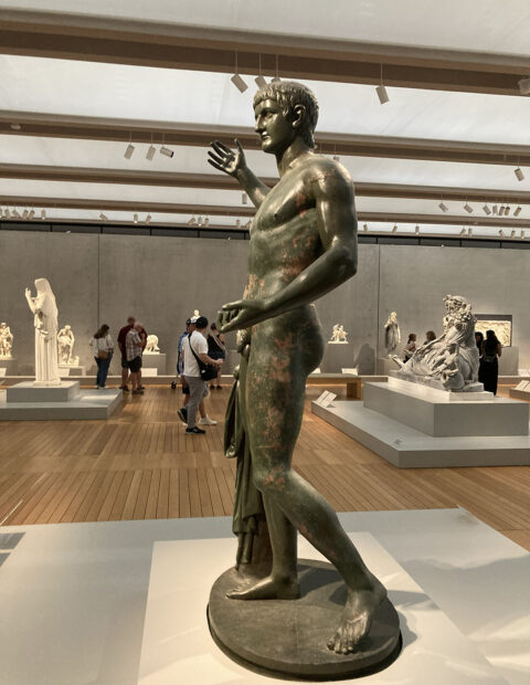 An installation image of a Roman bronze statue of a nude male figure, on view in a gallery with various other sculptures.