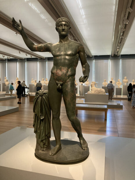 An installation image of a Roman bronze statue of a nude male figure, on view in a gallery with various other sculptures.