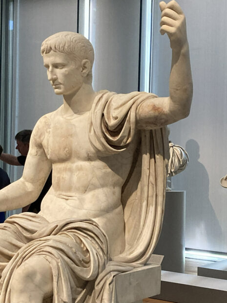 A photograph of a marble sculpture of a partially nude male figure. He is seated with his left arm raised as if holding a scepter. 