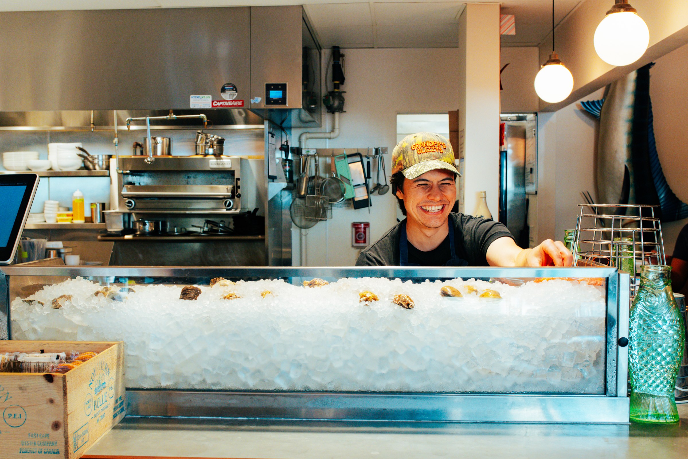A cook smiling as he digs into a clear box full of ice with oysters and shrimp on top.