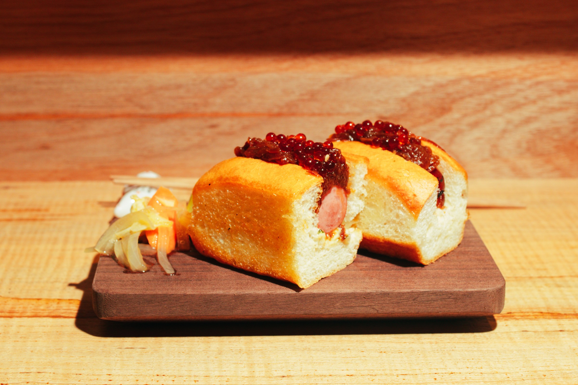 A hot dog topped with roe chopped in half sits on a wooden plate next to ginger.