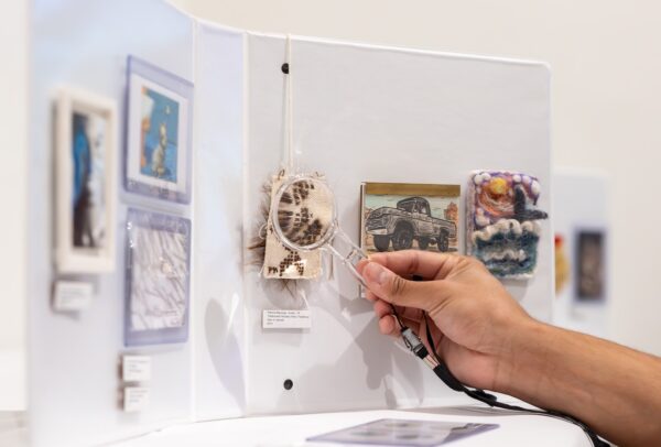 A hand holds a small magnifying glass in front of a miniature artwork.