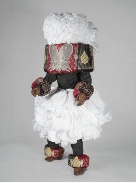 A complex African masquerade costume with white puffy top and skirt, exaggerated wrist and ankle cuffs, and a large cylindrical form around the head and neck area. 
