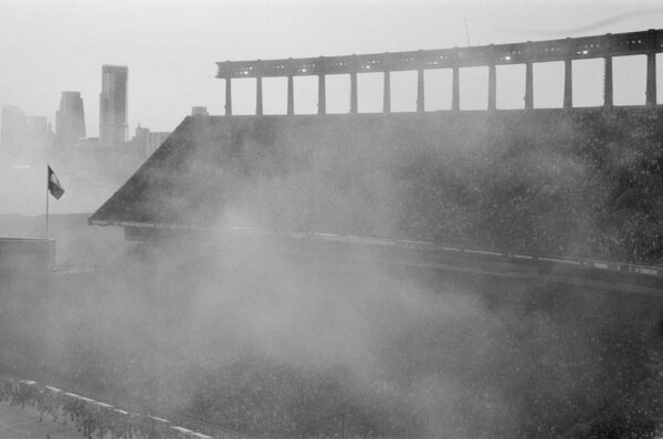A black and white photograph of smoke occluding a partial view of a stadium crowd.