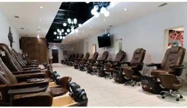 A long, modern nail salon interior featuring two rows of high-end, brown leather pedicure spa chairs lined up against white walls. The room is illuminated by elegant, spherical branch chandeliers and recessed lighting, creating a bright and clean atmosphe