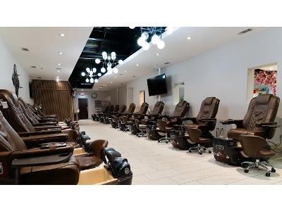 A long, modern nail salon interior featuring two rows of high-end, brown leather pedicure spa chairs lined up against white walls. The room is illuminated by elegant, spherical branch chandeliers and recessed lighting, creating a bright and clean atmosphe
