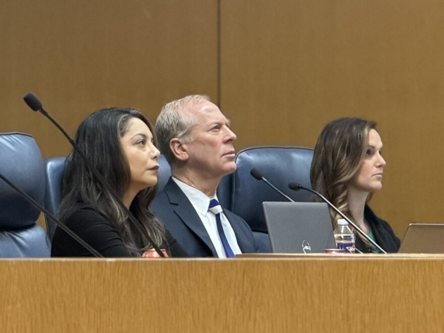 Three people in business attire sit behind a wooden desk and look forward. 
