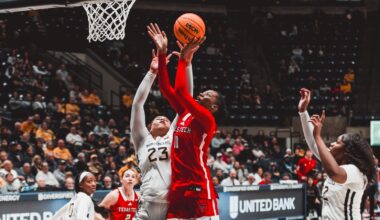 Texas Tech guard Bailey Maupin, wearing a red No. 20 jersey, looks up and drives a shot toward the basket as West Virginia defenders contest in front of a packed arena.