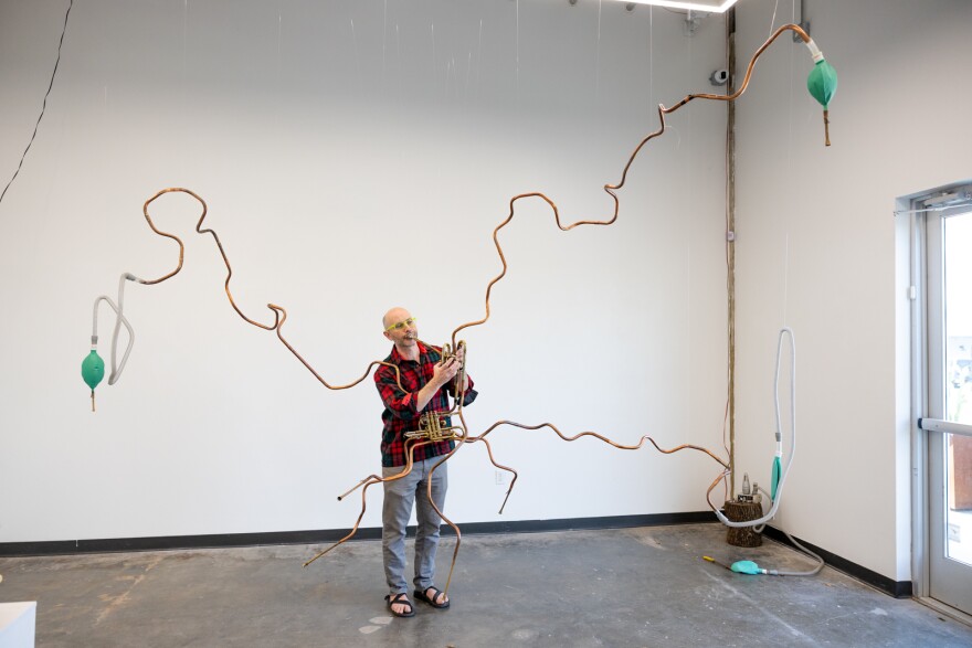 A man blows into an instrument similar to a trumpet that has piping extended into five separate lines that spread almost to the walls. 